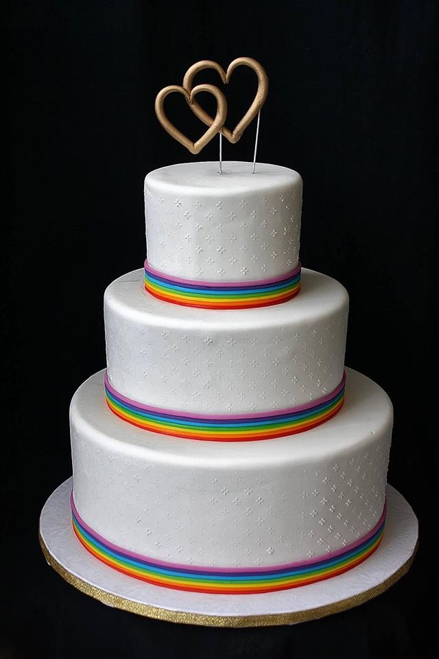 White three-tier wedding cake with rainbow ribbons and gold hearts topper